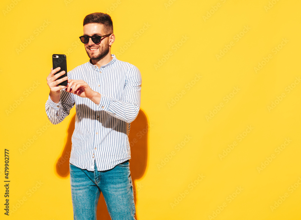 Handsome smiling model.Sexy stylish man dressed in shirt and jeans. Fashion hipster male posing near yellow wall in studio. Holding smartphone. Looking at cellphone screen. Using apps. Holding phone Handsome smiling model.Sexy stylish man dressed in shirt and jeans. Fashion hipster male posing near yellow wall in studio. Holding smartphone. Looking at cellphone screen. Using apps. Holding phone