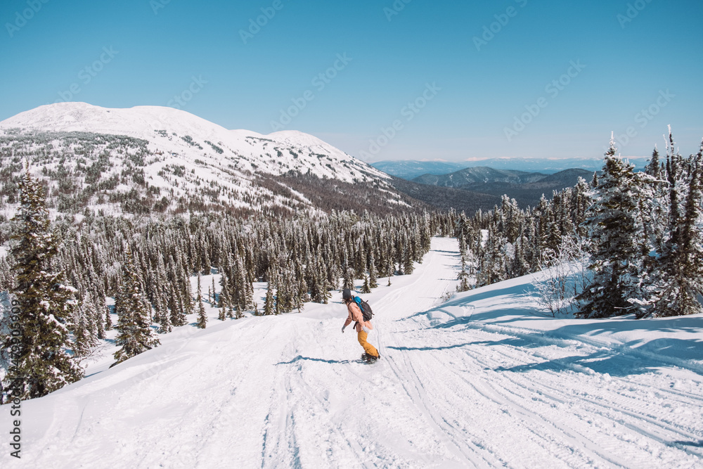 Russia. Sheregesh. Girl snowboarder in sun goggles in winter in sunny weather outdoors among the mountains and snow. Spring. Girl rides a snowboard in the distance, silhouette of a man among nature