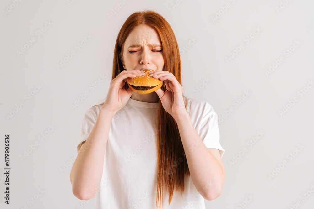 Studio shot of happy hungry young woman with closed eyes enjoying bite of appetizing delicious hamburger on white isolated background. Closeup front view of female eating tasty burger .