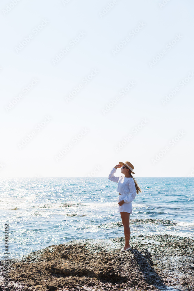An attractive young woman with long blond hair in a white summer suit stands on the seashore and enjoys the view of the ocean.