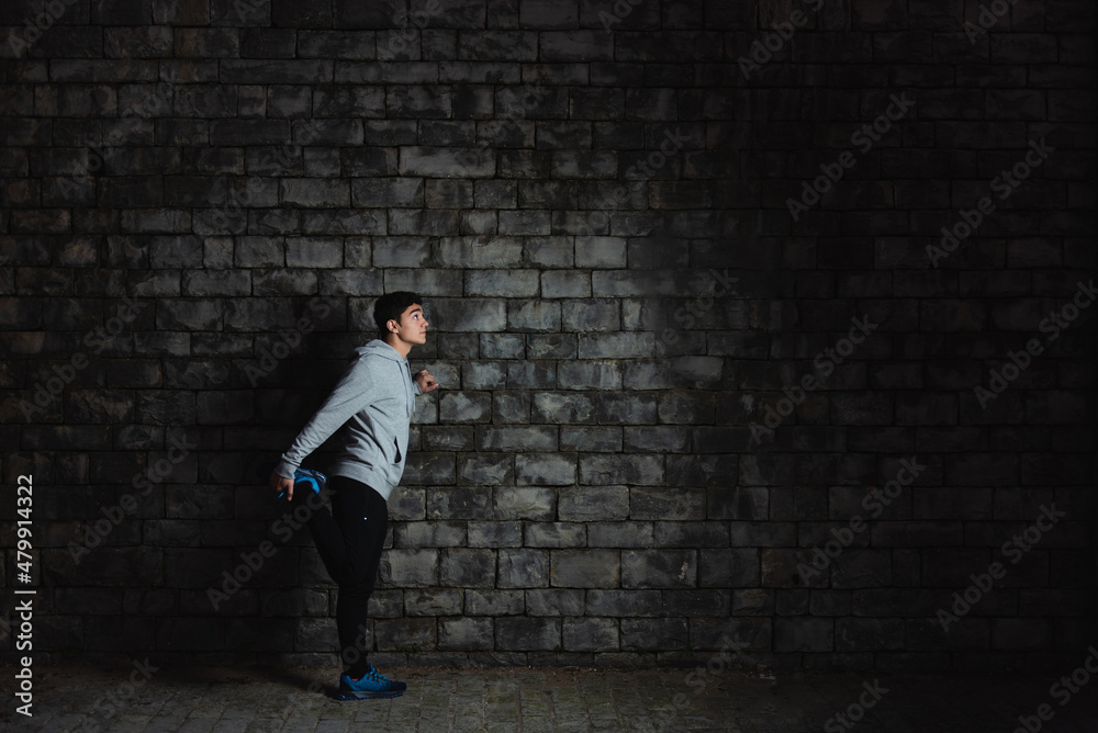 Hispanic teenager sportsman stretching against wall in the darkness. Hispanic teenager sportsman stretching against wall in the darkness.