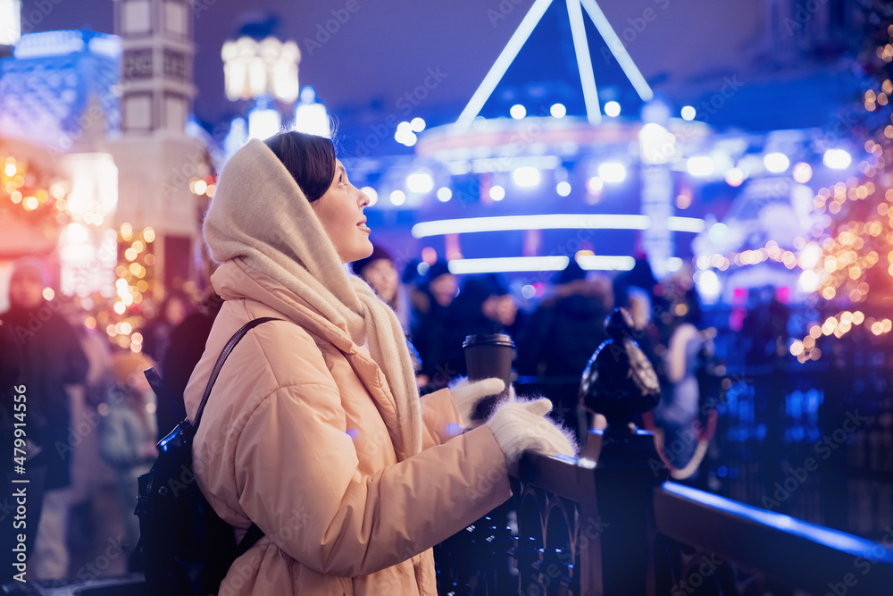 Happy joyful young woman with cup hot coffee walking down street on cold evening with bokeh light. Spending winter vacations in Red square, Moscow