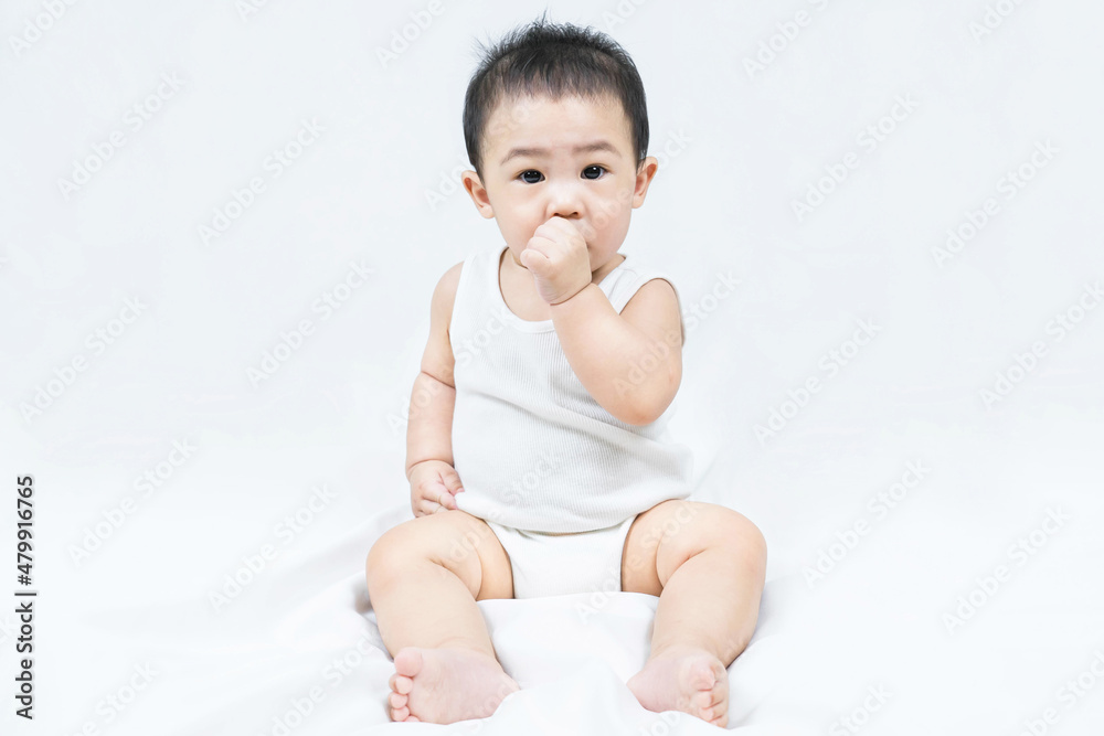 Adorable little seated baby boy in a white bodysuit sucking fingers looks at the camera on white background. Adorable little seated baby boy in a white bodysuit sucking fingers looks at the camera on white background.