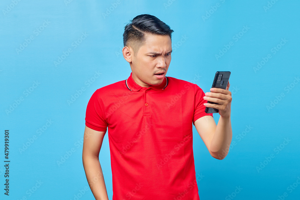 Portrait of angry young asian man looking at mobile phone, reading fake news isolated on blue background Portrait of angry young asian man looking at mobile phone, reading fake news isolated on blue background