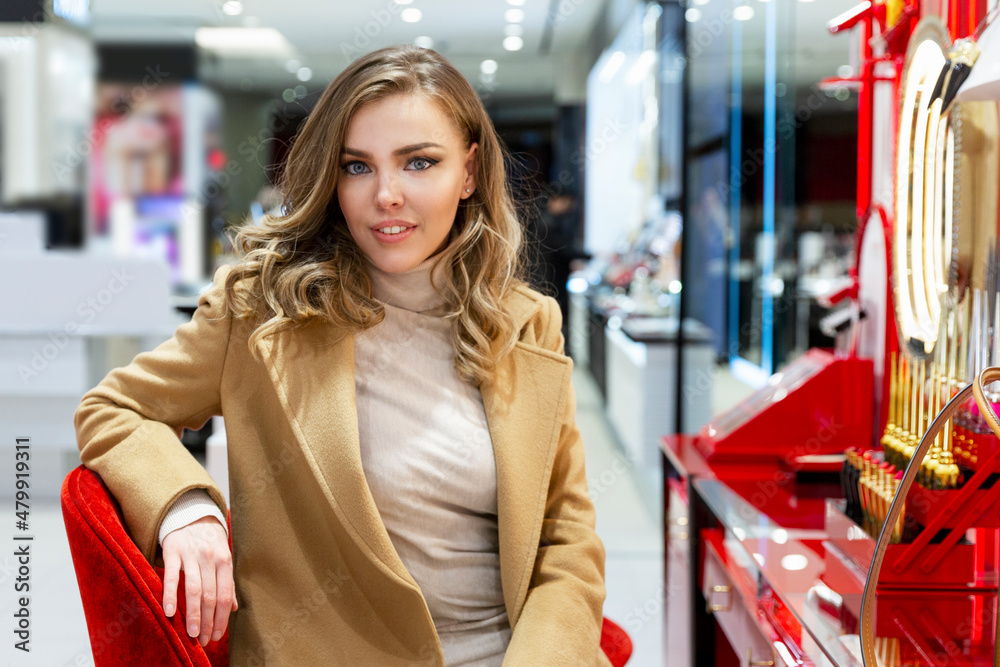 Beautiful woman sits in front of a mirror with cosmetics in a boutique. Smiling blonde in a beige coat is testing makeup. Beauty industry. Beautiful woman sits in front of a mirror with cosmetics in a boutique. Smiling blonde in a beige coat is testing makeup. Beauty industry.