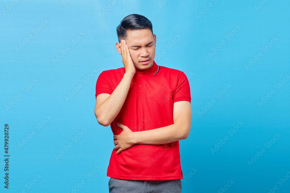 Portrait of handsome Asian young man in red shirt looking dizzy suffering from headache isolated on blue background