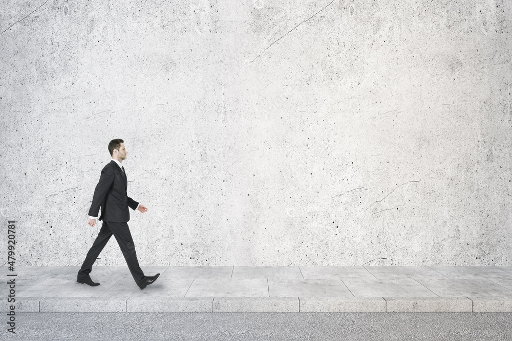 Side view of young businessman walking in concrete interior with mock up place on wall. Success concept. Side view of young businessman walking in concrete interior with mock up place on wall. Success concept.