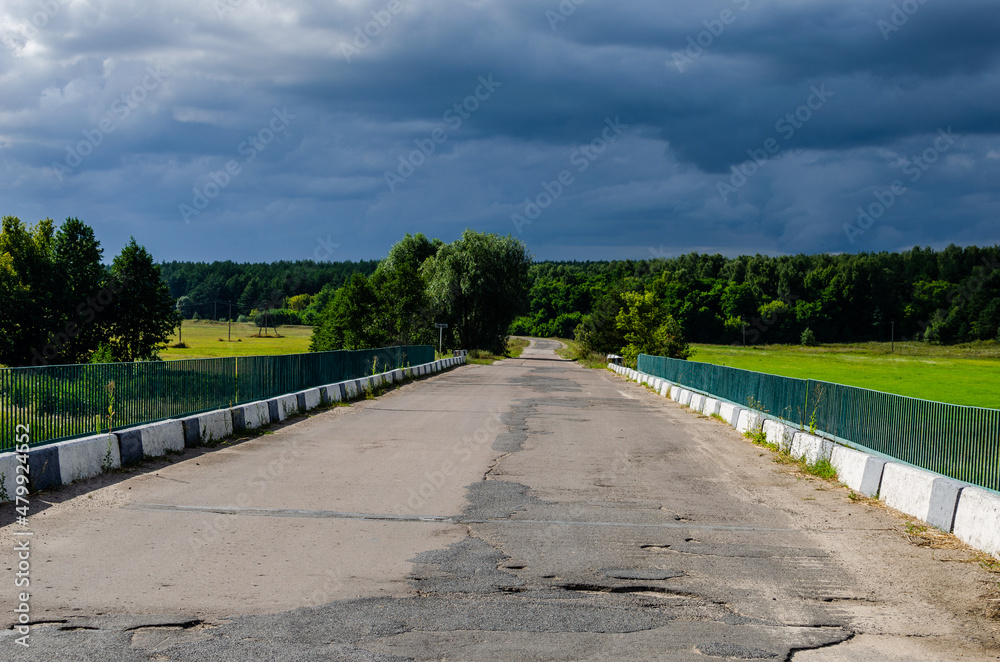 Thunderous sky over the field and forest. An asphalt road leading into the distance turns around a bend