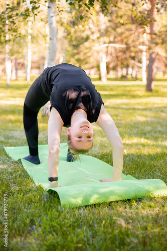 The girl is engaged in yoga, stands in a bridge pose on a sports mat in the park.
