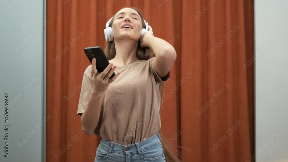Close-up cheerful woman singing and dancing in headphones on smartphone in room. Girl have fun moving listening to music relax in living room. Leisure concept at home Close-up cheerful woman singing and dancing in headphones on smartphone in room. Girl have fun moving listening to music relax in living room. Leisure concept at home