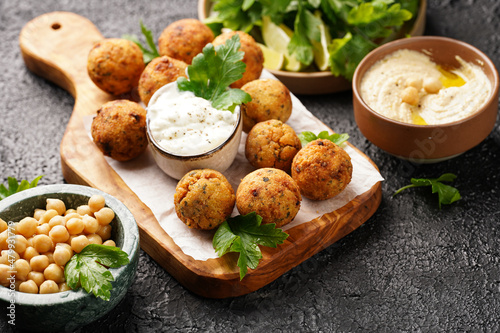 Traditional oriental chickpea deep fried falafel on a wooden board, tzatziki yoghurt sauce, hummus, fresh lime and green cilantro on black surface