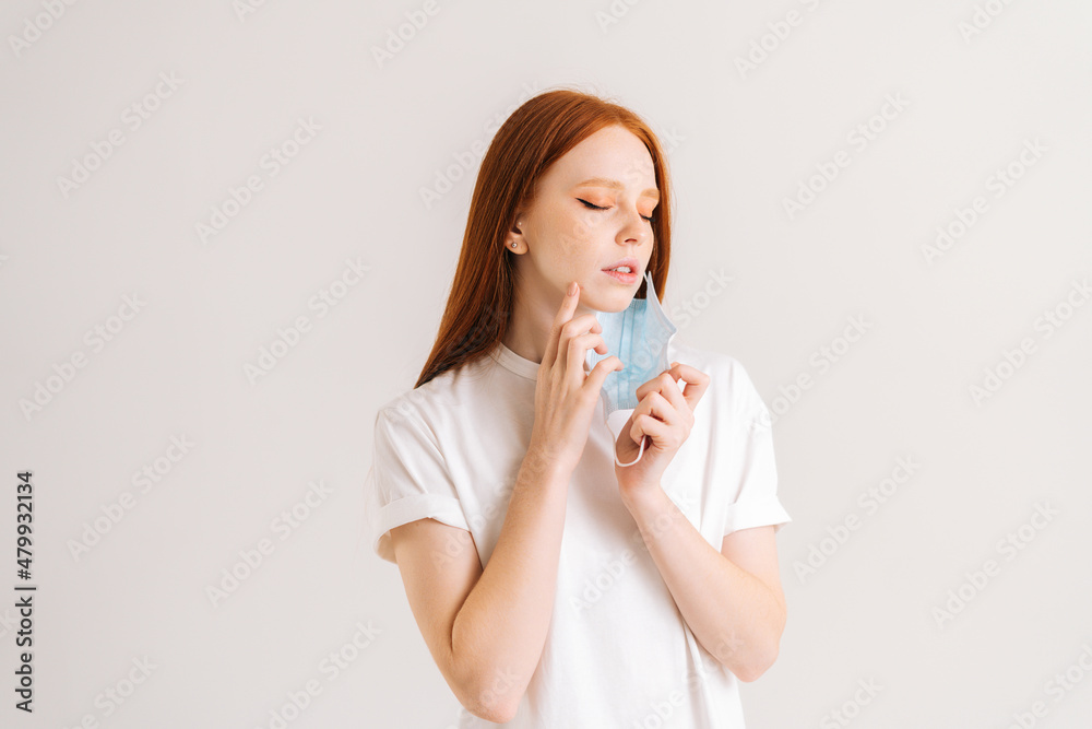 Studio portrait of pretty young woman wearing casual t-shirt taking off disposable protective face mask, standing on white isolated background. Flu epidemic, dust allergy, protection against virus Studio portrait of pretty young woman wearing casual t-shirt taking off disposable protective face mask, standing on white isolated background. Flu epidemic, dust allergy, protection against virus