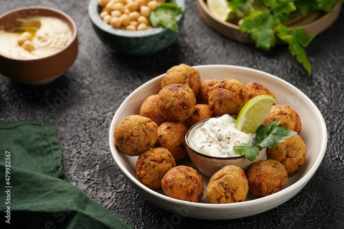 Traditional oriental chickpea deep fried falafel in a deep bowl with tzatziki yoghurt sauce, hummus, fresh lime and green cilantro on black surface