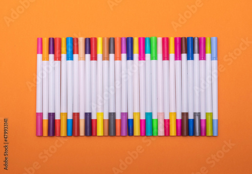A set of multi-colored markers (felt-tip pens) lies in an even row on an orange background.