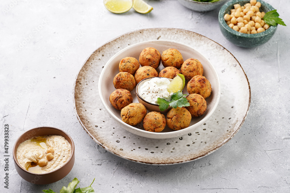 Traditional oriental chickpea deep fried falafel in a deep bowl with tzatziki yoghurt sauce, hummus, fresh lime and green cilantro on light grey concrete surface Traditional oriental chickpea deep fried falafel in a deep bowl with tzatziki yoghurt sauce, hummus, fresh lime and green cilantro on light grey concrete surface