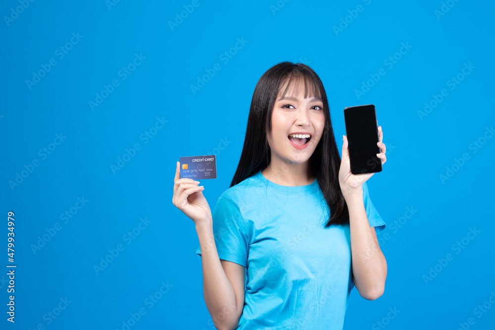 Portrait Asian happy young girl smiling cheerful and showing plastic credit card while holding mobile phone isolated on blue background Portrait Asian happy young girl smiling cheerful and showing plastic credit card while holding mobile phone isolated on blue background