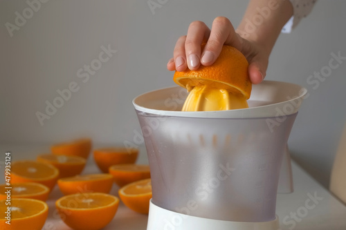 Woman is squeezing oranges using electric juicer in the kitchen, hand closeup. Healthy vegan ripe fruit juice, natural vitamins from food. Many halves of oranges on the kitchen table. photo