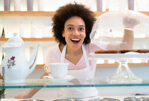 A beautiful mixed-race woman is smiling behind the stand of a pastry-gelato shop and showing the cookies under the glass bell.
