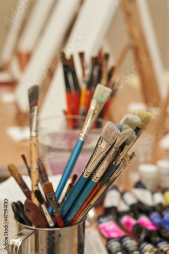 many paint brushes are in the glass in the foreground. in the background blank canvases in blur. artist's accessories