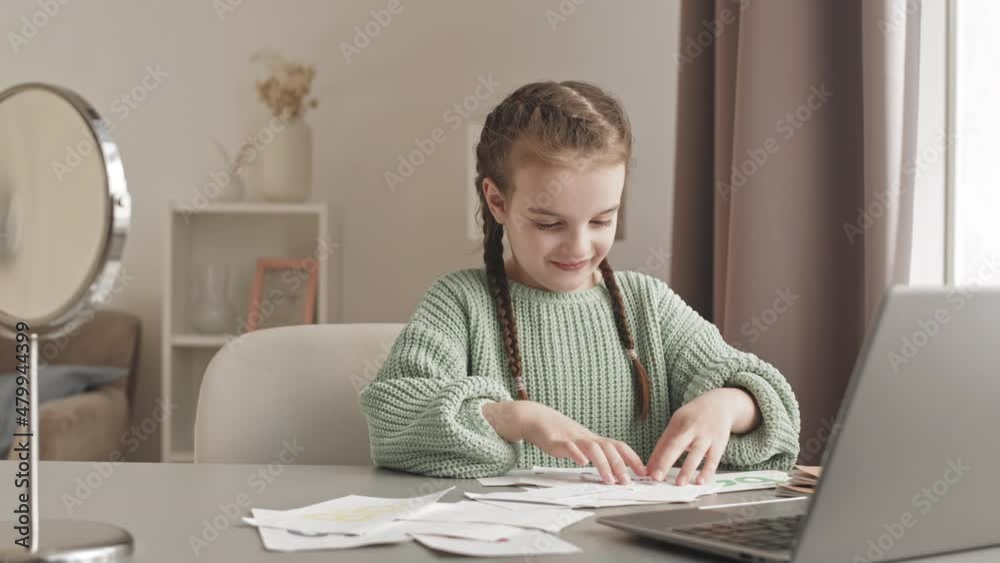 Waist up slowmo shot of smiling 5-year-old girl having online speech therapy lesson on laptop, showing printed cards with words and letters to teacher, sitting by desk at home Waist up slowmo shot of smiling 5-year-old girl having online speech therapy lesson on laptop, showing printed cards with words and letters to teacher, sitting by desk at home