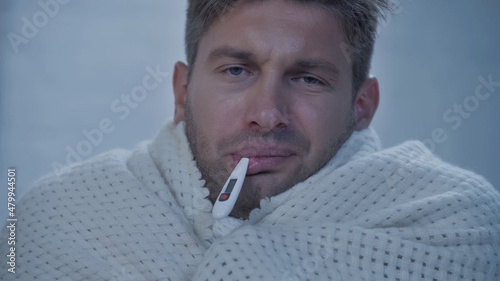 upset diseased man in blanket looking at camera while measuring temperature with thermometer in mouth.