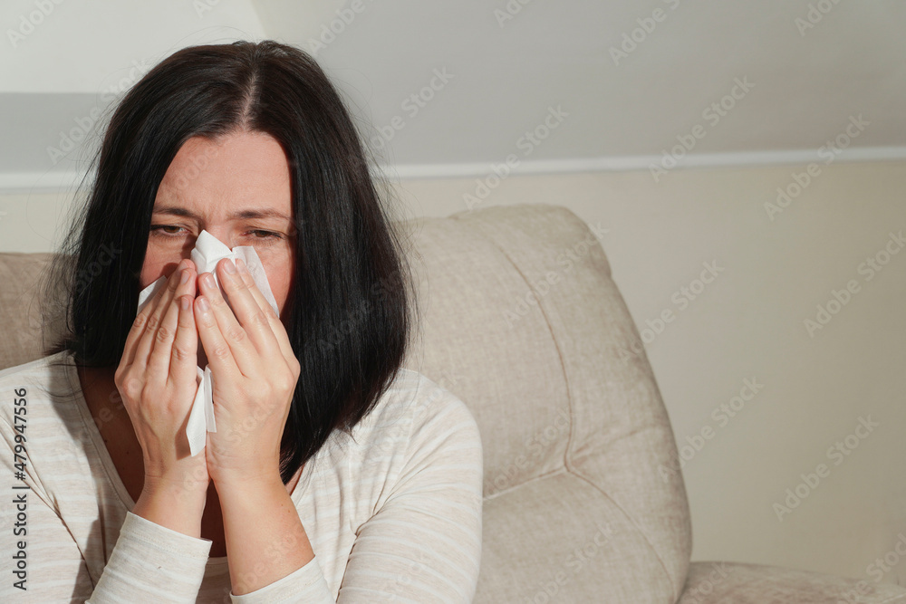Middle aged mature woman suffering from allergy rhinitis at home. Symptoms of cold or allergy. Woman sneezes in white tissue. Middle aged mature woman suffering from allergy rhinitis at home. Symptoms of cold or allergy. Woman sneezes in white tissue.