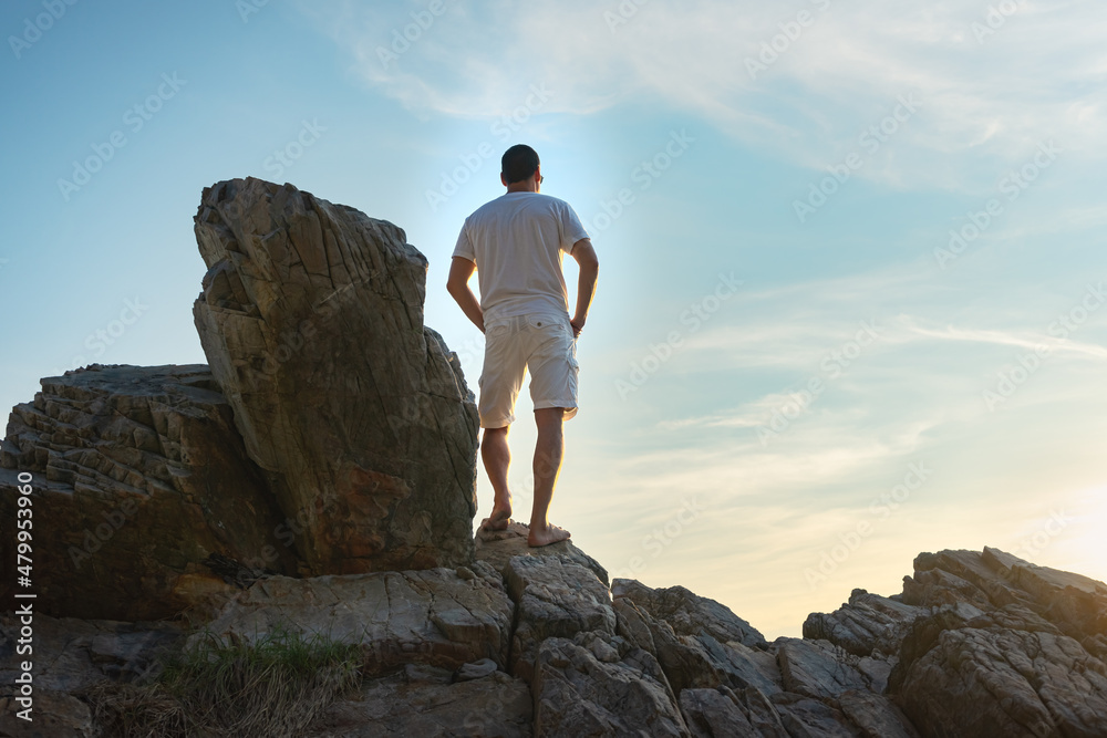 A man in a white t-shirt and shorts stands on top of a mountain and enjoys the sunset. Blue sky in the background.