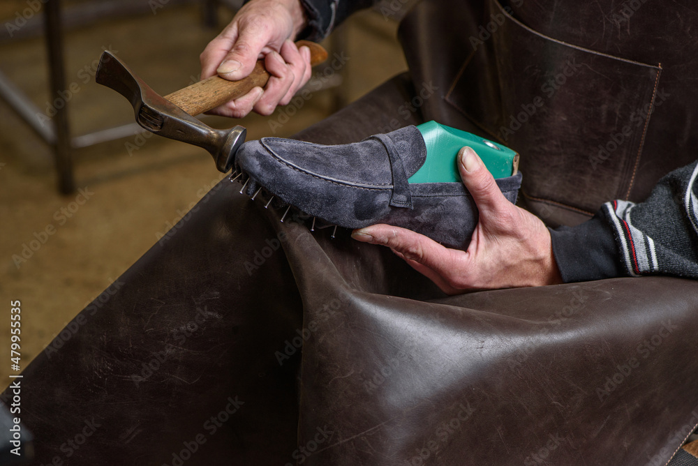 Shoemaker in leather apron making shoes in workshop. Male hands holding suede gray shoe on last and hammer. Shoemaking concept. Closeup.