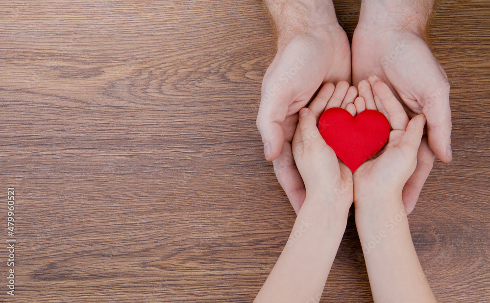 A red heart in the hands of a child and a man on a wooden background.The concept of love, family, health, charity