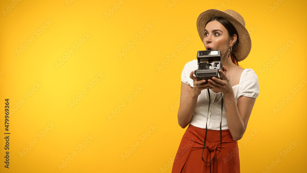 Obraz premium Excited tourist in straw hat holding vintage camera isolated on yellow.
