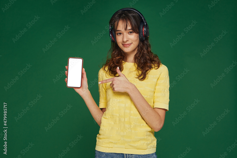 Stylish young girl in headphones point to mockup blank screen phone. Audiobooks, podcast music mobile apps advertising Stylish young girl in headphones point to mockup blank screen phone. Audiobooks, podcast music mobile apps advertising