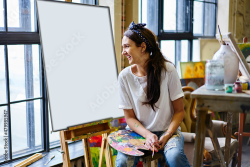 Happy Caucaisan woman with palette and paintbrush laughing during leisure pastime for creating sketch, joyful hipster girl with equipment for drawing enjoying own hobby during weekend in art studio