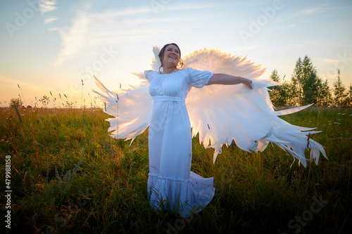 Plump chubby pleasant woman in white dres and angel wings in green grass field. Model girl posing outdoors on nature in a day or a evening
