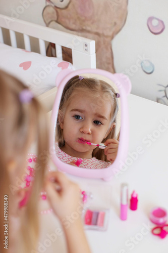 A little girl sits at a children's table in front of a mirror and paints her lips.