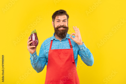 Happy man in apron showing OK gesture holding eggplant yellow background, greengrocer
