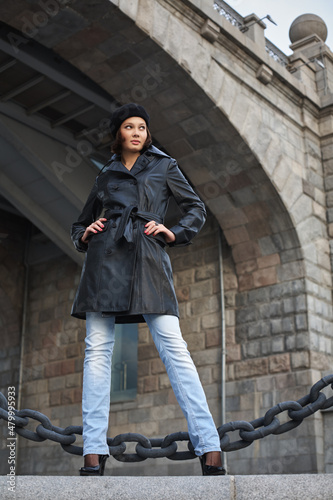 Beautiful young woman in a black leather coat and beret on the background of a stone bridge and a large chain. Full-length view.