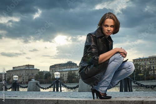 Beautiful young woman in a black leather coat sits on the stone steps of the embankment