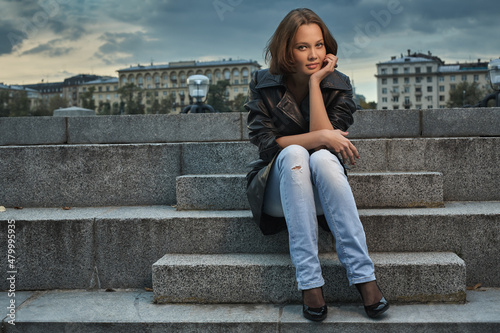 Beautiful young woman in a black leather coat sits on the stone steps of the embankment