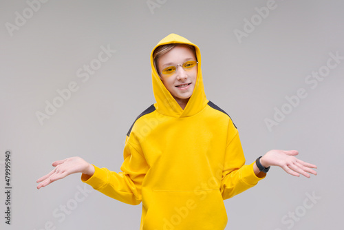 Funny curious young man in casual yellow sweatshirt, hood and yellow fashionable glasses posing cheerfully, caucasian teenager. Indoor studio shot isolated on light gray background