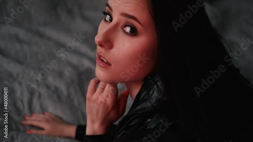 sexy confident brunette woman in red underwear and leather jacket on grey bed.