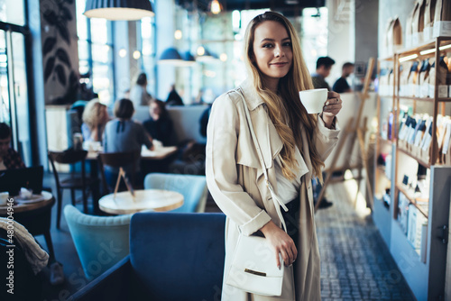 Fototapeta Young woman in trench coat having hot drink in cafe