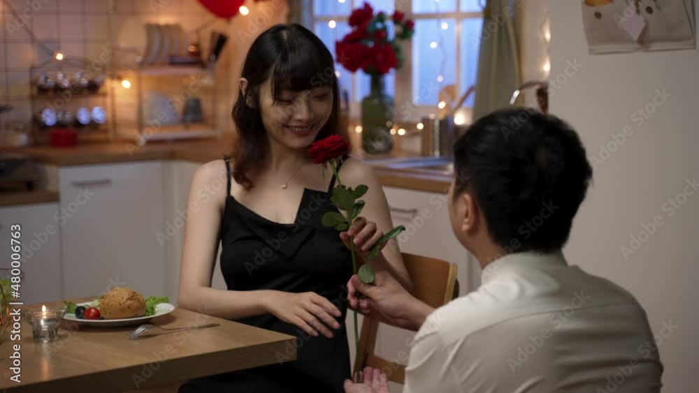 backside of asian male wearing suit surprising girlfriend with red rose behind. the woman smiling and nodding as her boyfriend gives flower on his knee during valentine’s night date at home backside of asian male wearing suit surprising girlfriend with red rose behind. the woman smiling and nodding as her boyfriend gives flower on his knee during valentine’s night date at home