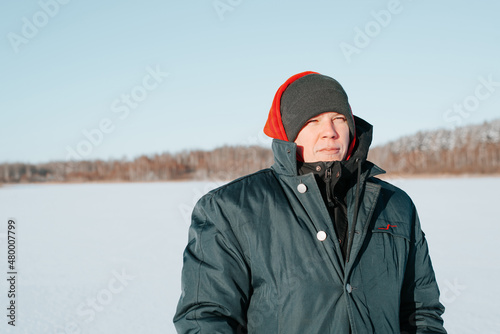 Portrait of caucasian man in winter inhat and hood, in warm jacket, standing in nature against background of snowy landscape and looking away