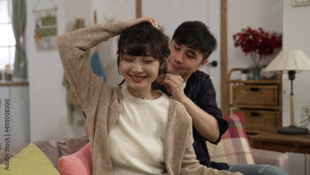 smiling asian woman holding up hair as her husband is putting on necklace for her, feeling happy about the beautiful gift. loving man hugging his wife on the sofa at home