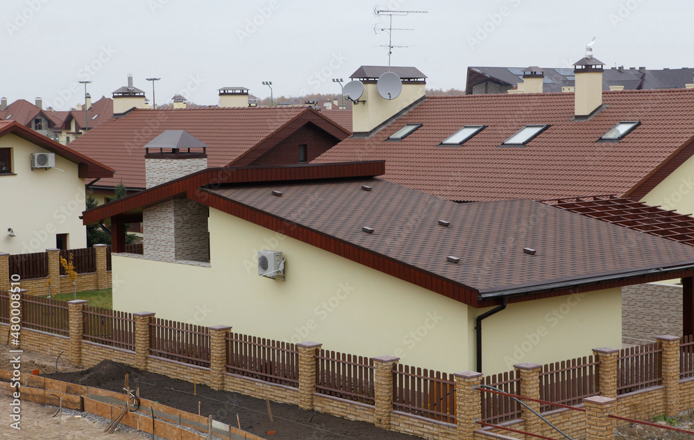 The view of many house roofs covered with bitumen, asphalt shingles, metal and clay tiles with skylights, ventilation caps, and chimneys. The construction of a cottage town with densely built houses.