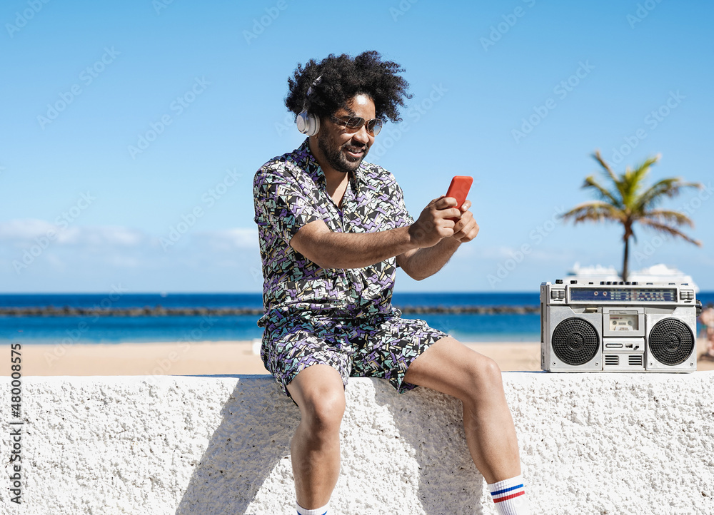 Obraz premium Cool latin boy sitting near the beach while listening music with vintage stereo and using mobile phone