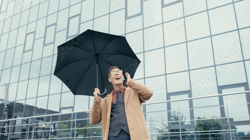 Happy businessman in coat and earphones holding umbrella on urban street.