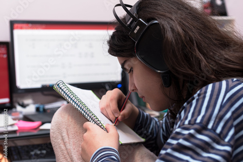 Transgender teenager with headphones and microphone studying in front of unfocused computer, listening to music while solving math homework in her notebook.