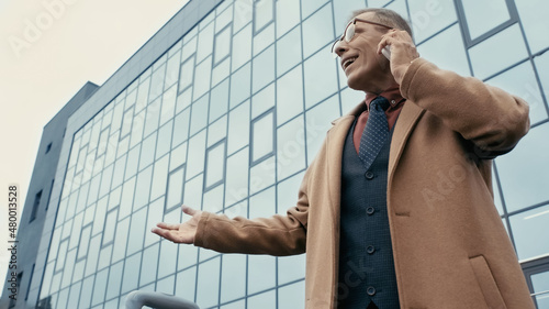 Low angle view of cheerful businessman in coat talking on smartphone on urban street.
