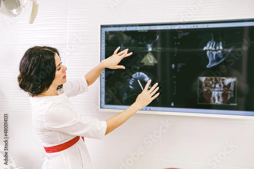 Serious dentist woman in white uniform and red belt showing attentively at the monitor with teeth snapshot.